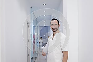 Portrait of a Young smiling bearded doctor holding a folder while leaning on white wall and looking to camera at a clinic corridor