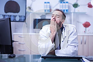 Young bearded doctor tired and yawning sitting at the office in his hospital cabinet