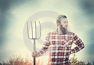 Young bearded farmer in red checkered shirt with old pitchfork sky nature backgrund, toned photo