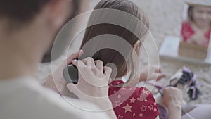 Young bearded father brushing the hair of his little girl while the child combing her toy sitting on floor on fluffy