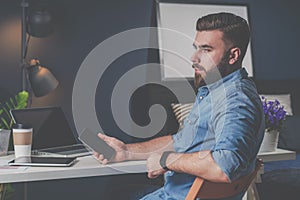 Young bearded freelancer man in denim shirt is sitting at home at table, holding mobile phone in his hand and thinking.