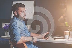 Young bearded freelancer man in denim shirt is sitting at home at table, holding mobile phone in his hand and thinking.