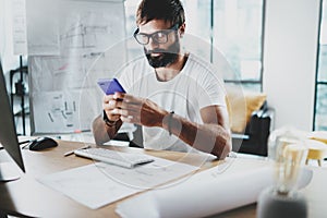 Young bearded graphic designer wearing eye glasses and working at modern loft studio-office.Man using smartphone.Blurred