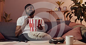 Young bearded guy watching uninteresting film while sitting on sofa at home. Handsome African man in cap eating popcorn