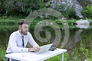 Young, bearded guy on worktable in summer, using laptop. man working, studying, surfing web. remote workplace in nature