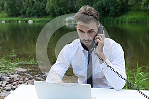 Young, bearded guy on worktable in summer, using laptop, talking on phone. man working, studying or surfing web in park