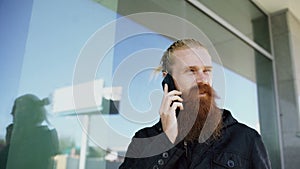 Young bearded hipster man concentrated talking on phone on citystreet and have conversation near office building