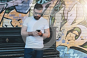 Young bearded hipster man dressed in white t-shirt and sunglasses is stands against wall with graffiti