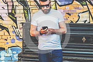 Young bearded hipster man dressed in white t-shirt and sunglasses is stands against wall with graffiti