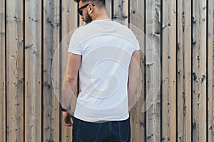Young bearded hipster man dressed in white t-shirt and sunglasses is stands outdoor against wood wall. Mock up.