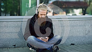 Young bearded hipster man with headphones sitting on road and using smartphone for listen to music and internet surfing
