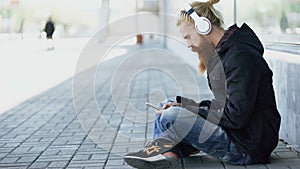 Young bearded hipster man with headphones sitting on road and using smartphone for listen to music and internet surfing