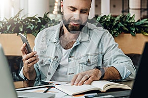 Young bearded hipster man sits in cafe in front of computers, reads notes in notebook while holding smartphone in hand.