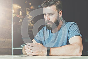 Young bearded hipster man sits at a table in a cafe and uses a smartphone. Guy is chatting, working, blogging, learning