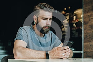 Young bearded hipster man sits at a table in office and uses a smartphone. Guy is chatting, blogging, learning online