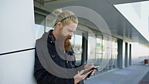 Young bearded hipster man using smartphone fo internet surfing near offfice building