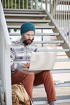 Young bearded hipster student using computer outdoors