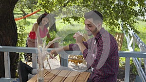 Young bearded lazy man drinking tea in the foreground while angry attractive girl in retro clothes planting flowers in