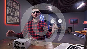 Young bearded male gives five to camera having box of power supply on desk.