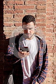Young bearded male leaning on a bricked wall while using a smartphone