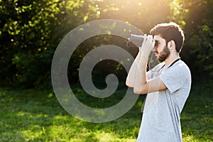 Young bearded male looking through binoculars while standing at green field, observing something. Handsome young guy having expedi