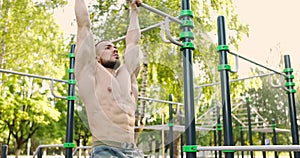 Young bearded man with a bare torso doing leg lifting on a horizontal bar on sports ground