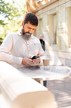 Young bearded man in business shirt attire standing in relaxing outdoor cafe drinking coffee while using mobile phone