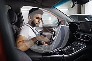 Young bearded man car technician mechanic sitting inside red car repairing problem of engine