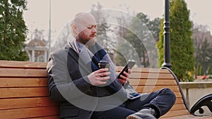 Young bearded man in casual sitting on bench in park drinking cup of coffee and using his mobile phone