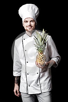 Young bearded man chef In white uniform holds Fresh pineapple on black background