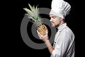 Young bearded man chef In white uniform holds Fresh pineapple on black background