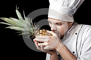Young bearded man chef In white uniform holds Fresh pineapple on black background
