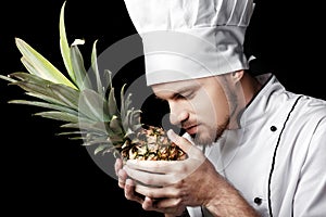 Young bearded man chef In white uniform holds Fresh pineapple on black background