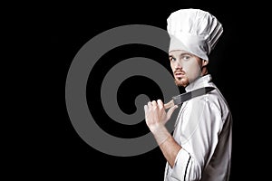 Young bearded man chef In white uniform holds knife on black background