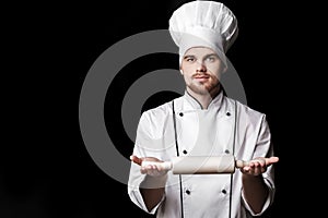 Young bearded man chef In white uniform holds rolling pin on black background