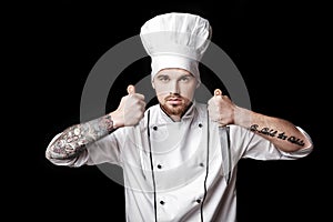 Young bearded man chef In white uniform holds Two knives on black background