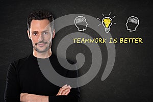 Young bearded man with crossed arms, heads with brains, lightbulb-idea and text `Teamwork is better` on a blackboard background.