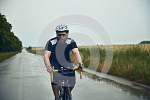 Young bearded man, cyclist in helmet, glasses and uniform riding bike on wet road in cloudy chill evening. Forest ride