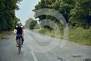 Young bearded man, cyclist in helmet, glasses and uniform riding bike on wet road in cloudy chill evening. Forest ride