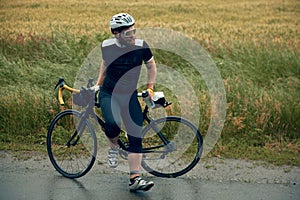Young bearded man, cyclist in uniform, helmet and glasses sitting on bicycle by road around field background. Stop for