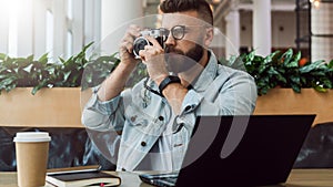 Young bearded man in denim jacket sits in coffee shop at table in front of laptop and takes instant photo on camera.