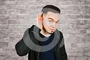 Young bearded man eavesdrops with the hand at ear on brick wall background