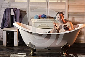 Young bearded man with foam on face shaving with razor in bathtub. Health beauty and skin care concept. Guy in bathroom.