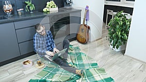 Young bearded man in glasses talking by phone and using laptop computer sitting in kitchen at home. Male speaking on the phone wit