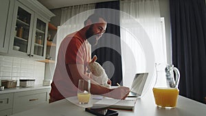 A young bearded man in a good mood stands sideways in the kitchen, uses a laptop to searching and typing information