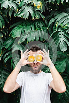 Young bearded man holding slices of orange tangerine in front of his eyes, surprised