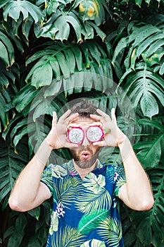 Young bearded man holding slices of Pitaya dragon fruit in front of his eyes, surprised