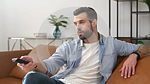 A young bearded man holding tv remote control, watching TV