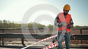 Young bearded man inspector in an orange vest and protective helmet with tablet