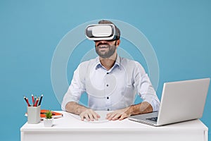 Young bearded man in light shirt sit and work at white desk with pc laptop isolated on pastel blue background in studio